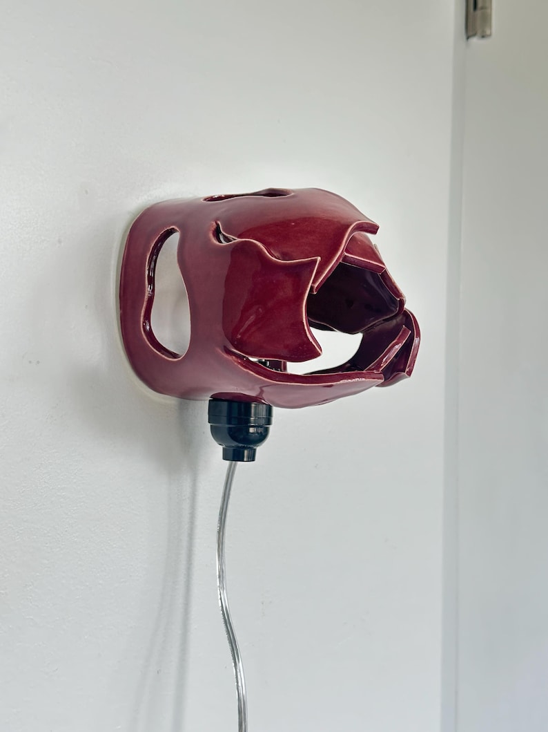 May include: A burgundy ceramic wall sconce with an unusual, organic form. It features a bulb socket and a silver cord. The sconce is mounted on a white wall.