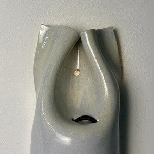 May include: A wall-mounted ceramic vessel with a glossy, light gray glaze and a smooth, undulating surface. The top features two folded, petal-like openings that converge toward a central cavity containing a small, dark, curved metal hook.