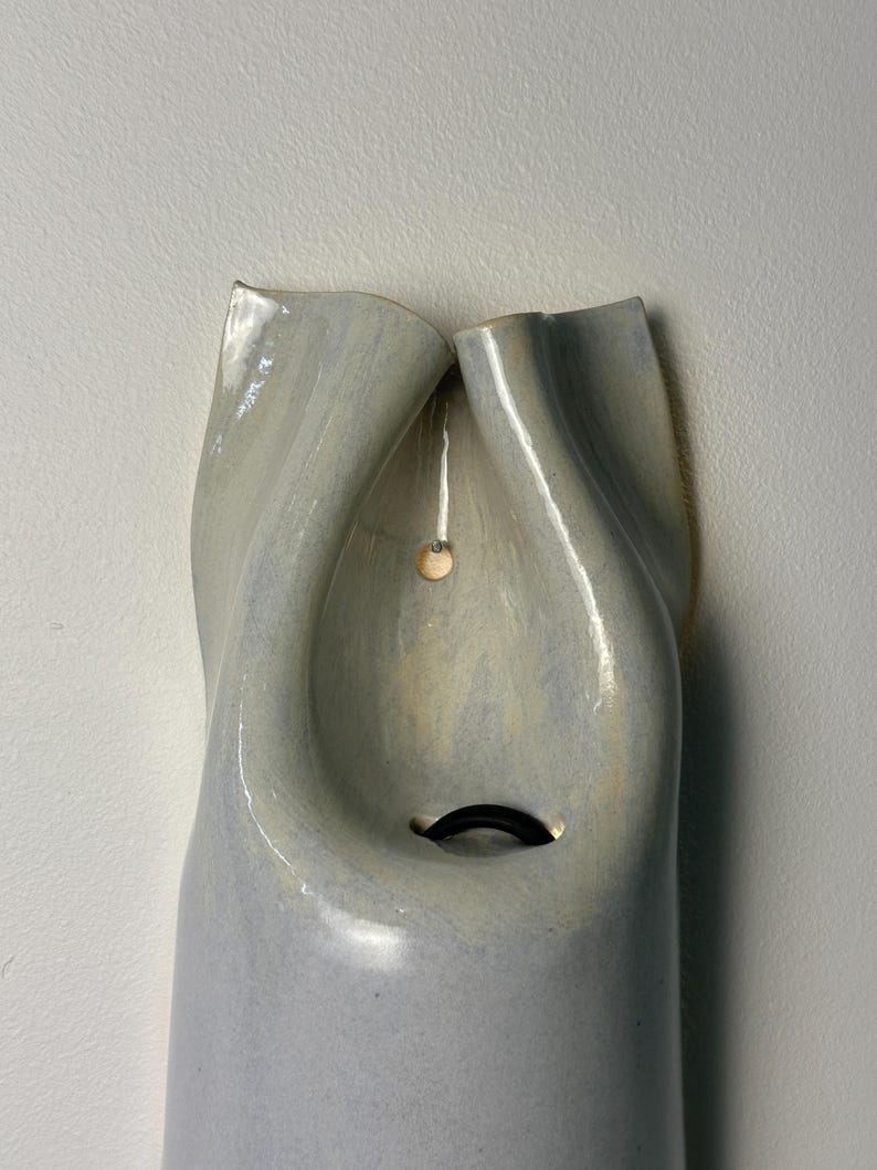 May include: A wall-mounted ceramic vessel with a glossy, light gray glaze and a smooth, undulating surface. The top features two folded, petal-like openings that converge toward a central cavity containing a small, dark, curved metal hook.