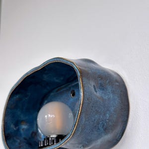 May include: A blue ceramic wall sconce with a bulb. The sconce has a unique, organic shape with a textured surface and a gold-colored rim. The bulb is round and emits a soft light.