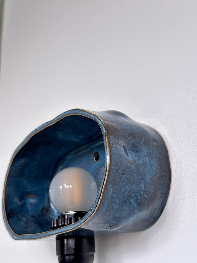 May include: A blue ceramic wall sconce with a bulb. The sconce has a unique, organic shape with a textured surface and a gold-colored rim. The bulb is round and emits a soft light.