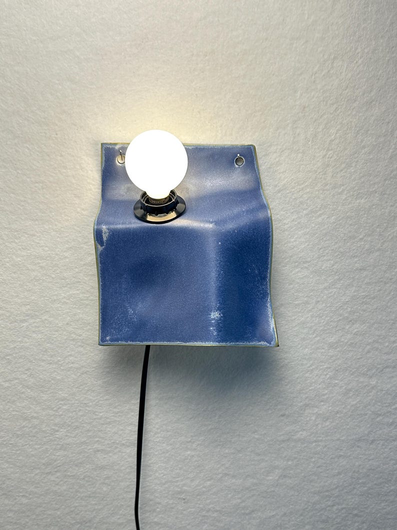 May include: A wall-mounted light fixture featuring a square, blue ceramic panel with a curved, folded shape. A single round, glowing white light bulb is mounted in the upper left section of the ceramic piece, which includes two small metal eyelets.
