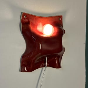 K&ouml;nnte beinhalten: Eine Wandleuchte aus Keramik mit einer gl&auml;nzenden, tiefroten Glasur und einer wellenf&ouml;rmigen, organischen Form. Das Objekt verf&uuml;gt &uuml;ber eine zentrale kreisf&ouml;rmige Aussparung f&uuml;r eine leuchtende wei&szlig;e Kugelbirne und zwei Befestigungsl&ouml;cher.