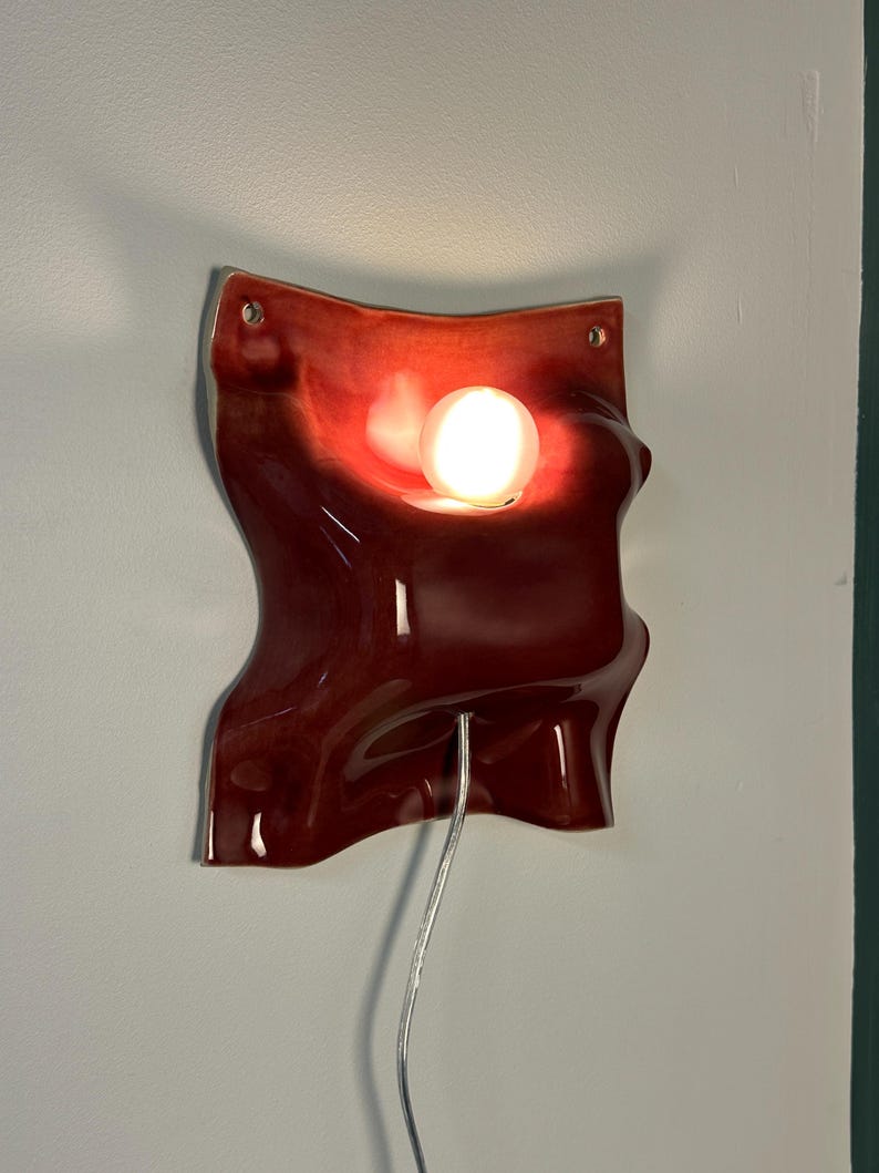 K&ouml;nnte beinhalten: Eine Wandleuchte aus Keramik mit einer gl&auml;nzenden, tiefroten Glasur und einer wellenf&ouml;rmigen, organischen Form. Das Objekt verf&uuml;gt &uuml;ber eine zentrale kreisf&ouml;rmige Aussparung f&uuml;r eine leuchtende wei&szlig;e Kugelbirne und zwei Befestigungsl&ouml;cher.