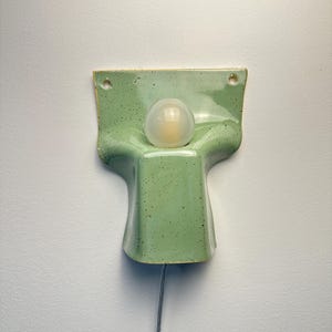 May include: A wall-mounted ceramic light fitting featuring a glossy, light green speckled glaze. The structure has a flared, rectangular top section with two mounting holes and a central, rounded protrusion holding a single frosted glass bulb.