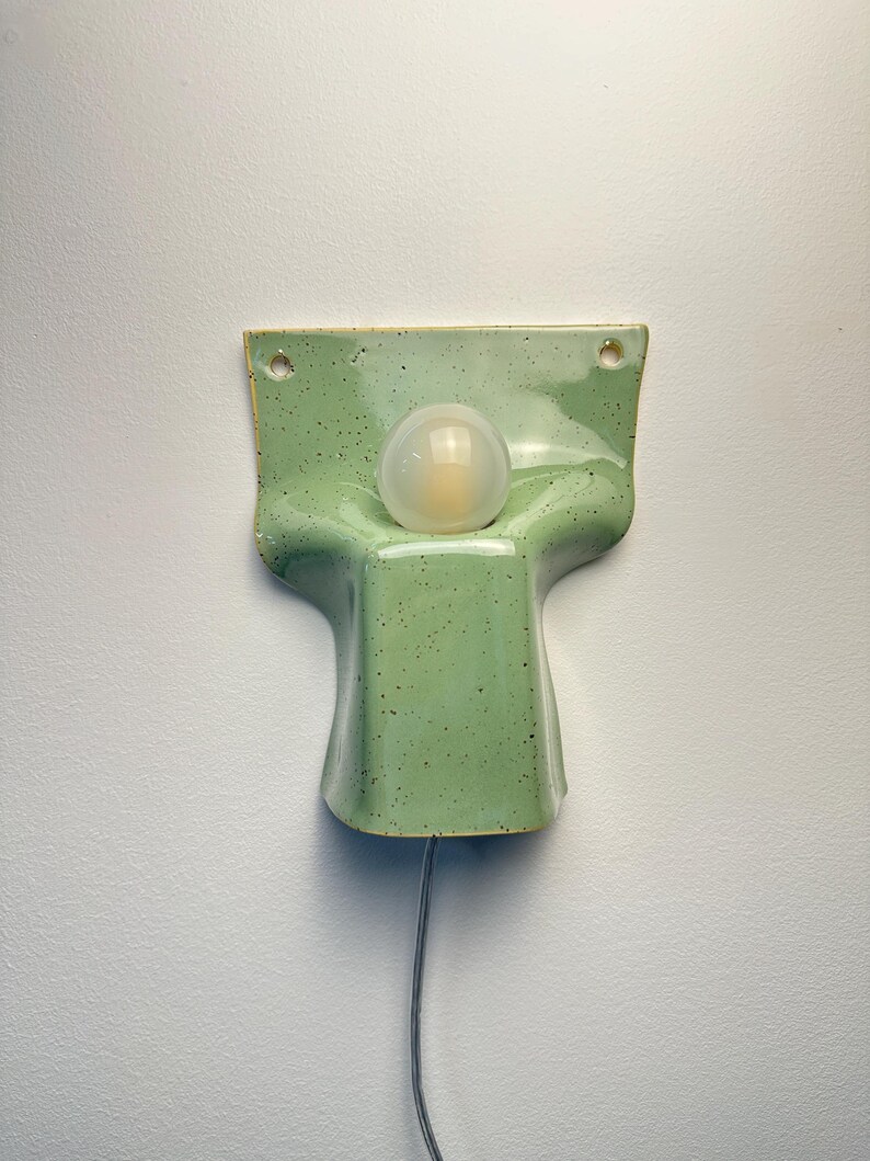 May include: A wall-mounted ceramic light fitting featuring a glossy, light green speckled glaze. The structure has a flared, rectangular top section with two mounting holes and a central, rounded protrusion holding a single frosted glass bulb.
