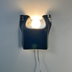 May include: A wall-mounted ceramic sconce with a glossy dark blue glaze and a central cutout. A round, glowing white light bulb is positioned within the recessed area. A thin, clear electrical cord hangs from the bottom of the fixture.