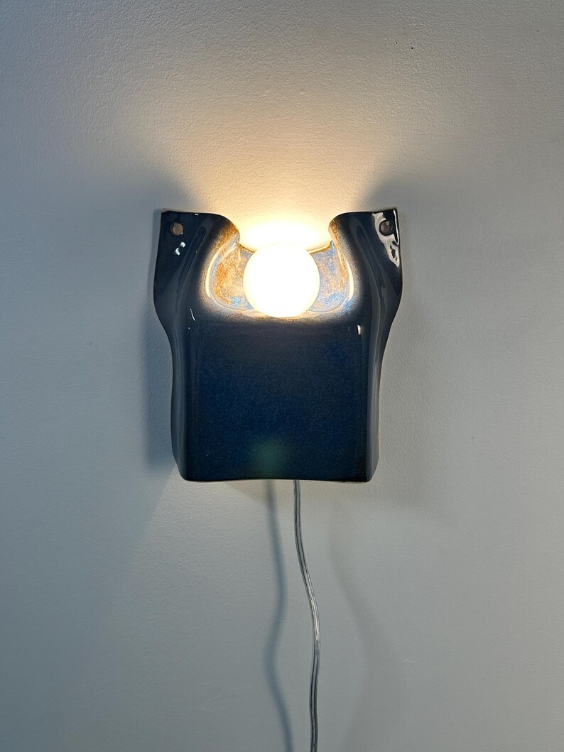 May include: A wall-mounted ceramic sconce with a glossy dark blue glaze and a central cutout. A round, glowing white light bulb is positioned within the recessed area. A thin, clear electrical cord hangs from the bottom of the fixture.
