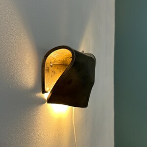 May include: A wall-mounted sconce featuring a curved, dark metallic exterior shell with a reflective gold-toned interior. The fixture emits a warm, glowing light from the bottom and sides, casting soft illumination against the wall surface.