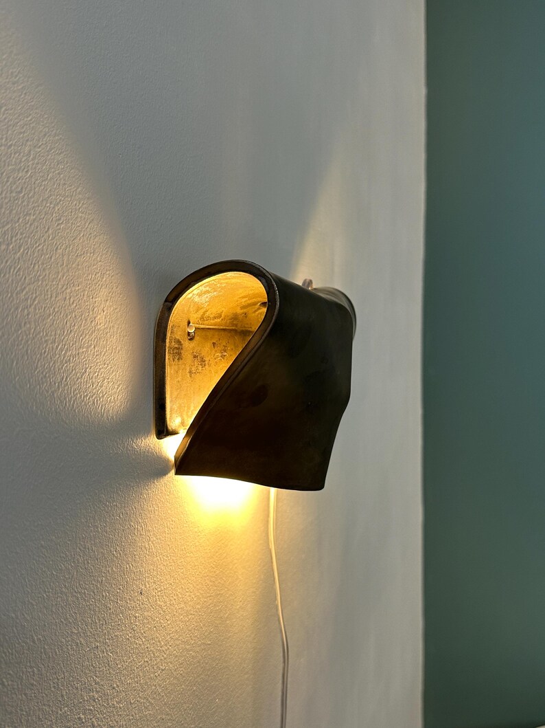 May include: A wall-mounted sconce featuring a curved, dark metallic exterior shell with a reflective gold-toned interior. The fixture emits a warm, glowing light from the bottom and sides, casting soft illumination against the wall surface.