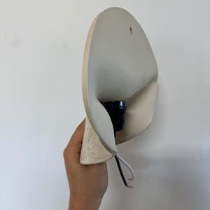 May include: A wall sconce featuring a curved, off-white ceramic shade with a smooth finish. The fixture includes a black central socket for a light bulb and a short black and white electrical cord extending from the base for installation.