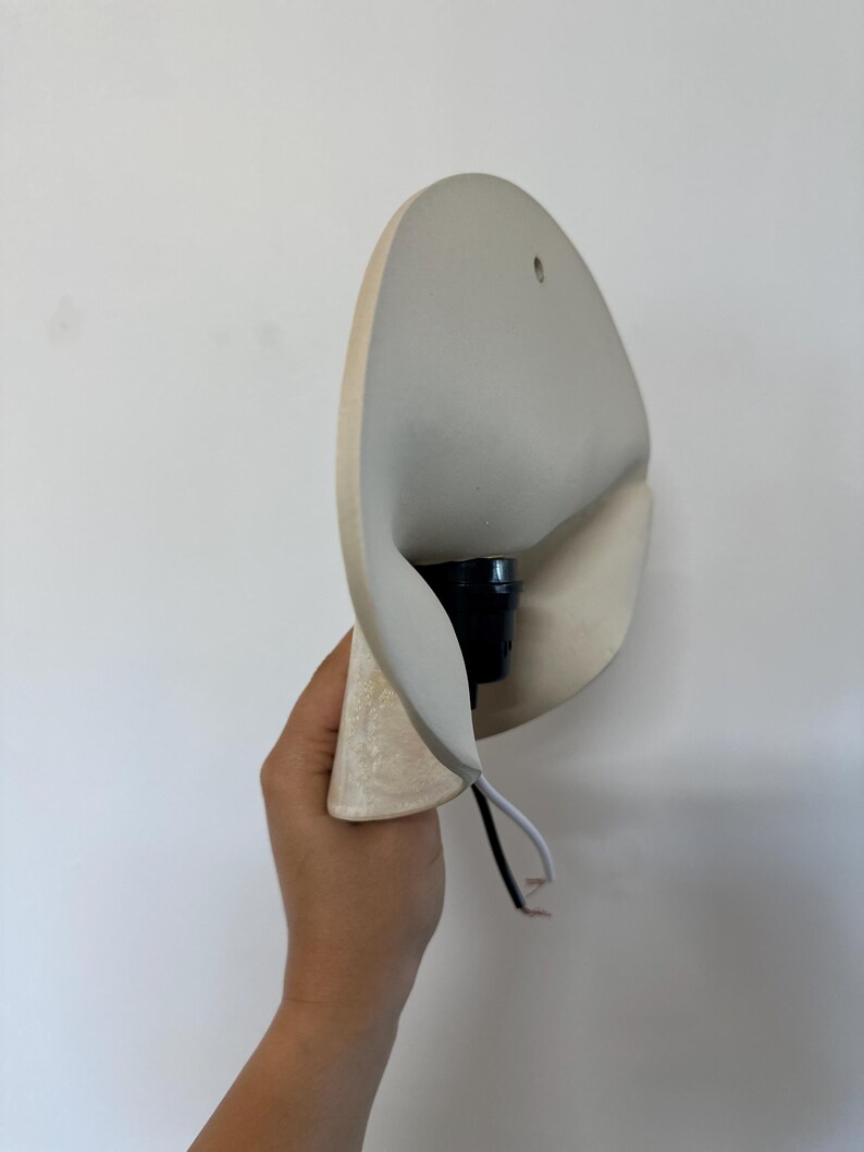 May include: A wall sconce featuring a curved, off-white ceramic shade with a smooth finish. The fixture includes a black central socket for a light bulb and a short black and white electrical cord extending from the base for installation.