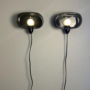 May include: Two oval wall sconces with a dark, smoky glass finish. Each sconce has a round, illuminated light bulb and a black cord. The sconces are mounted on a light grey wall, offering a modern lighting solution for any room.