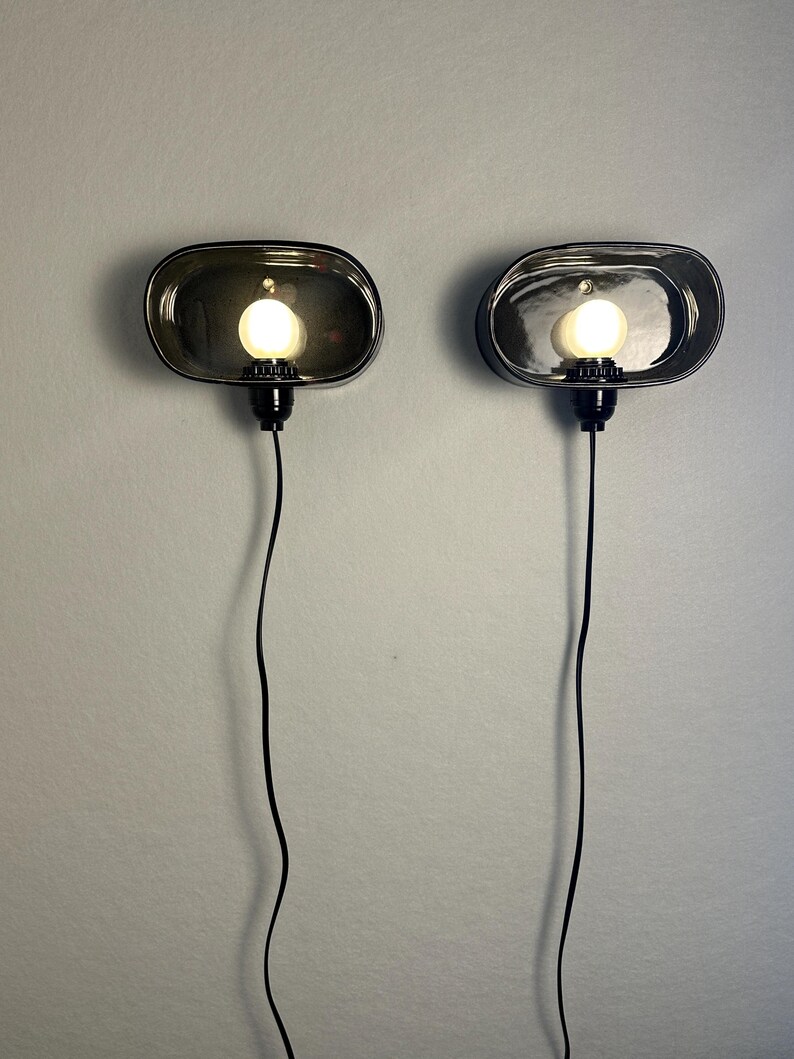 May include: Two oval wall sconces with a dark, smoky glass finish. Each sconce has a round, illuminated light bulb and a black cord. The sconces are mounted on a light grey wall, offering a modern lighting solution for any room.