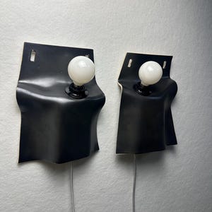 May include: Two wall-mounted lamps featuring a matte black, curved, sheet-like structure with a rectangular top section containing a mounting slot. Each lamp includes a single white spherical light bulb and a thin gray power cord extending downward.