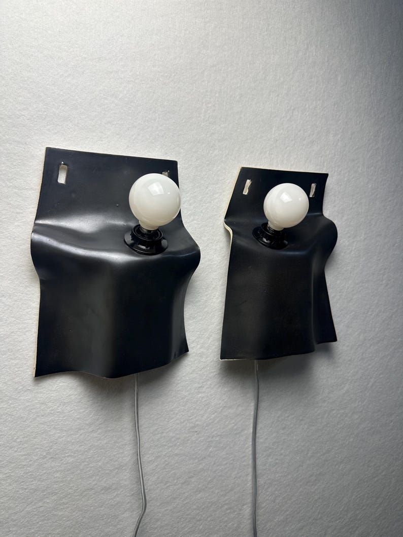 May include: Two wall-mounted lamps featuring a matte black, curved, sheet-like structure with a rectangular top section containing a mounting slot. Each lamp includes a single white spherical light bulb and a thin gray power cord extending downward.