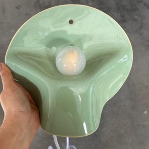 May include: A wall-mounted light fixture featuring a glossy, sage green ceramic shade with a fluid, organic, wavy shape. A single, round, frosted white light bulb is centered in the middle of the shade, which includes a small hole at the top.