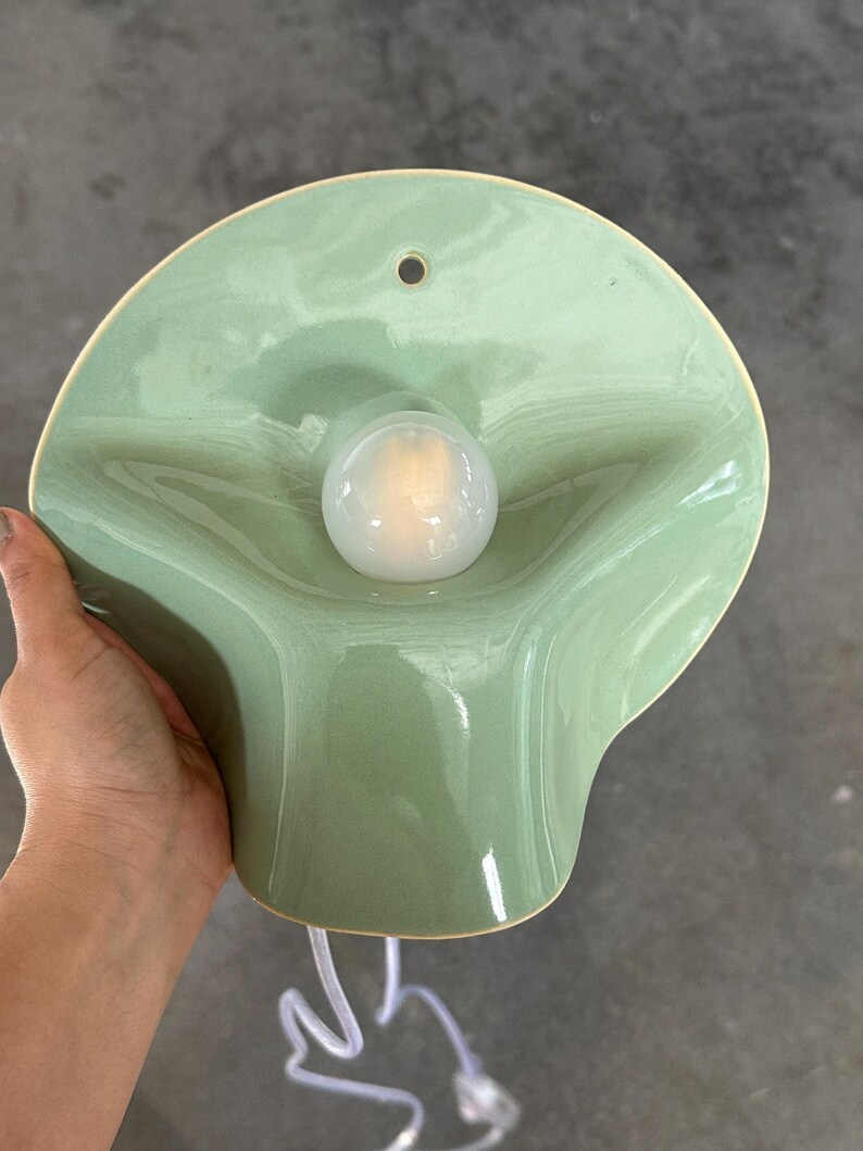 May include: A wall-mounted light fixture featuring a glossy, sage green ceramic shade with a fluid, organic, wavy shape. A single, round, frosted white light bulb is centered in the middle of the shade, which includes a small hole at the top.