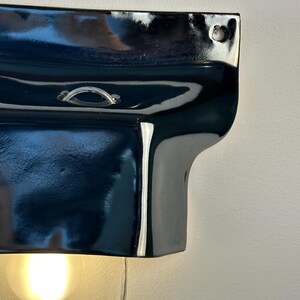 May include: A glossy black ceramic wall sconce with a curved, organic shape. The fixture features a small metal loop at the top center and a circular mounting hole in the upper right corner. A single warm-toned light bulb glows from underneath.