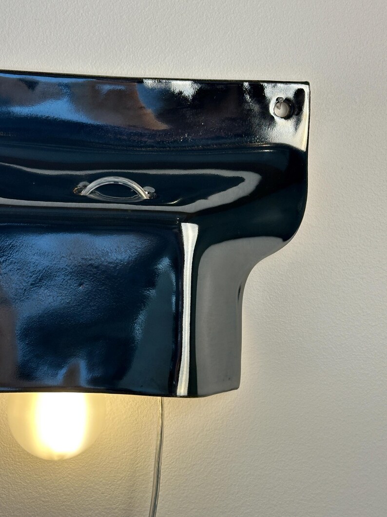 May include: A glossy black ceramic wall sconce with a curved, organic shape. The fixture features a small metal loop at the top center and a circular mounting hole in the upper right corner. A single warm-toned light bulb glows from underneath.
