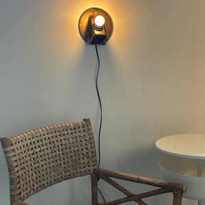 May include: A wall-mounted sconce with a circular metallic backplate and a single glowing light bulb. The fixture features a dark, angled housing and is connected to a thin black power cord that hangs vertically down the wall.