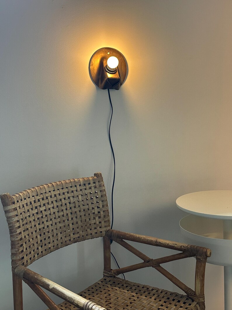 May include: A wall-mounted sconce with a circular metallic backplate and a single glowing light bulb. The fixture features a dark, angled housing and is connected to a thin black power cord that hangs vertically down the wall.