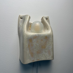 May include: A wall-mounted ceramic light fixture with a glossy, off-white and beige mottled glaze. The structure features a curved, U-shaped top section holding a single round, frosted glass light bulb, with two small holes for mounting screws.
