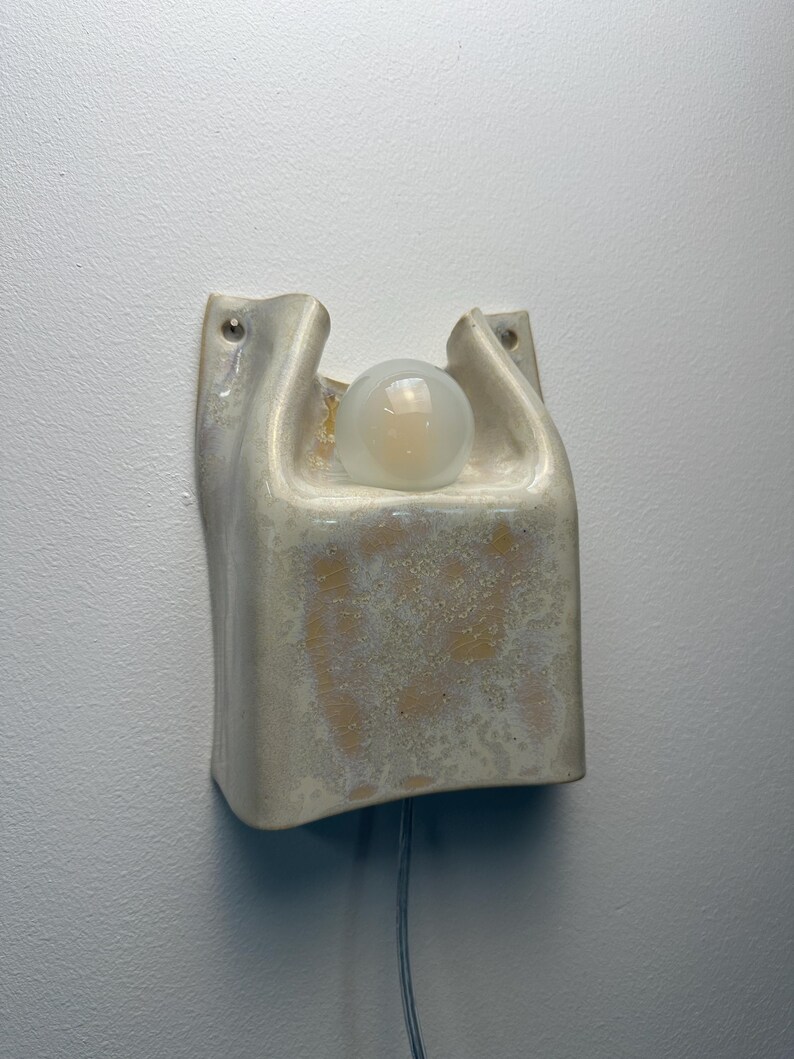 May include: A wall-mounted ceramic light fixture with a glossy, off-white and beige mottled glaze. The structure features a curved, U-shaped top section holding a single round, frosted glass light bulb, with two small holes for mounting screws.