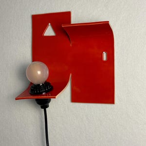 May include: A contemporary wall light with a vibrant red geometric shape. It has a small, spherical light bulb with a black base and a black cable. The design incorporates a triangular cut-out and a curved edge.