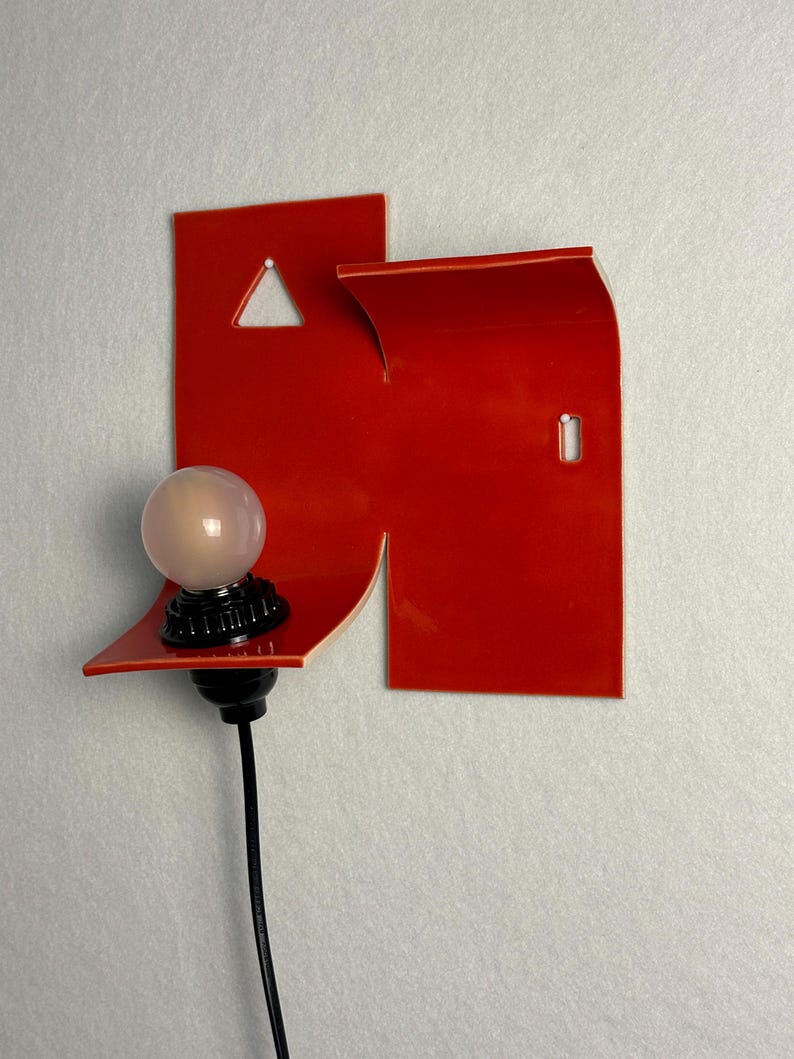 May include: A contemporary wall light with a vibrant red geometric shape. It has a small, spherical light bulb with a black base and a black cable. The design incorporates a triangular cut-out and a curved edge.