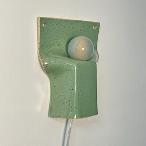 May include: A wall-mounted ceramic sconce in a light green speckled glaze. The fixture features a rectangular backplate with two mounting holes and a curved, protruding front section that holds a single, round, frosted white glass light bulb.