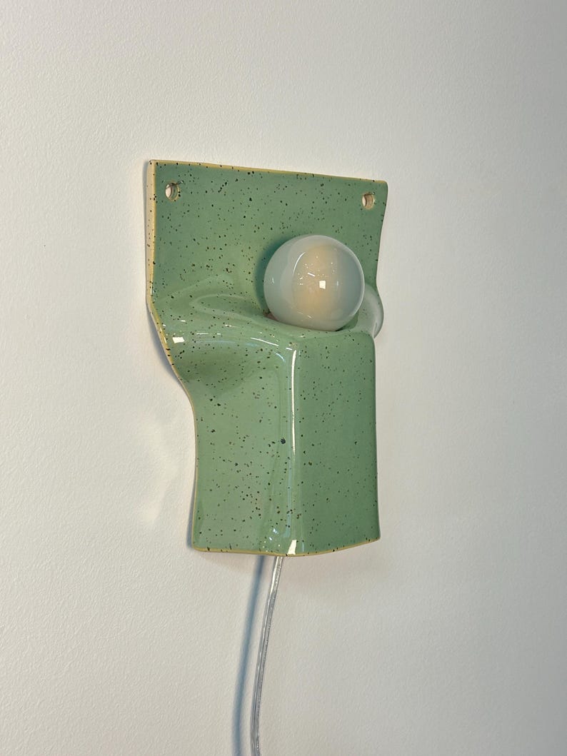May include: A wall-mounted ceramic sconce in a light green speckled glaze. The fixture features a rectangular backplate with two mounting holes and a curved, protruding front section that holds a single, round, frosted white glass light bulb.