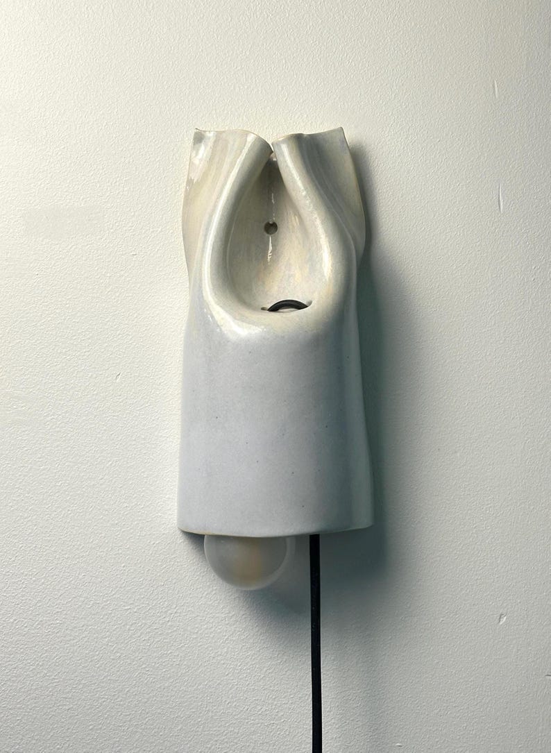 May include: A wall-mounted light fixture made of white ceramic with a smooth, glossy finish. The structure features a folded, organic shape with a central opening, a small circular mounting hole, and a single round light bulb extending from the bottom.
