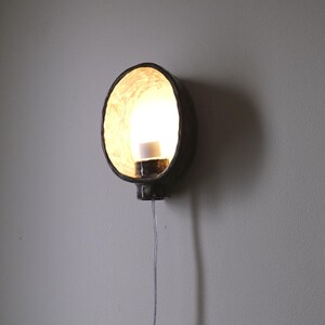 May include: A dark, round wall sconce with a bright light emanating from within. The sconce is mounted on a gray wall and has a cord hanging down. The interior of the sconce has a textured, light-colored surface.