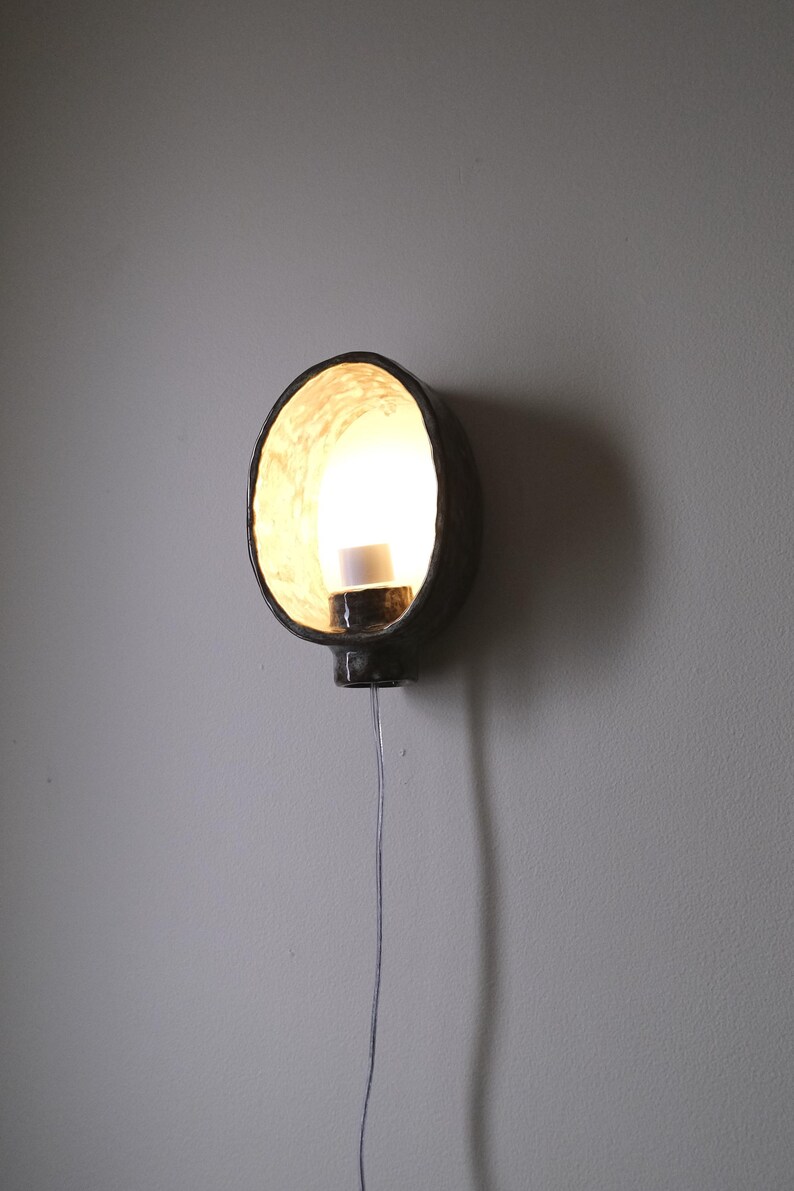 May include: A dark, round wall sconce with a bright light emanating from within. The sconce is mounted on a gray wall and has a cord hanging down. The interior of the sconce has a textured, light-colored surface.