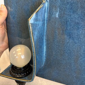 May include: A blue ceramic wall sconce with a gold trim. The sconce features a curved design and holds a round, frosted light bulb. The base of the bulb is black and has a decorative pattern. The sconce is mounted on a blue wall.