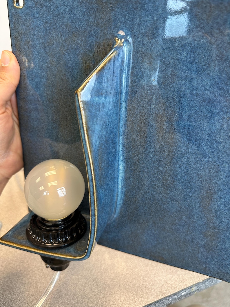 May include: A blue ceramic wall sconce with a gold trim. The sconce features a curved design and holds a round, frosted light bulb. The base of the bulb is black and has a decorative pattern. The sconce is mounted on a blue wall.