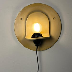Pu&ograve; includere: Un'applique circolare color giallo senape con una sporgenza centrale ad arco che sostiene una lampadina smerigliata luminosa. L'apparecchio include un portalampada nero e un cavo di alimentazione nero, con due piccoli fori di montaggio.