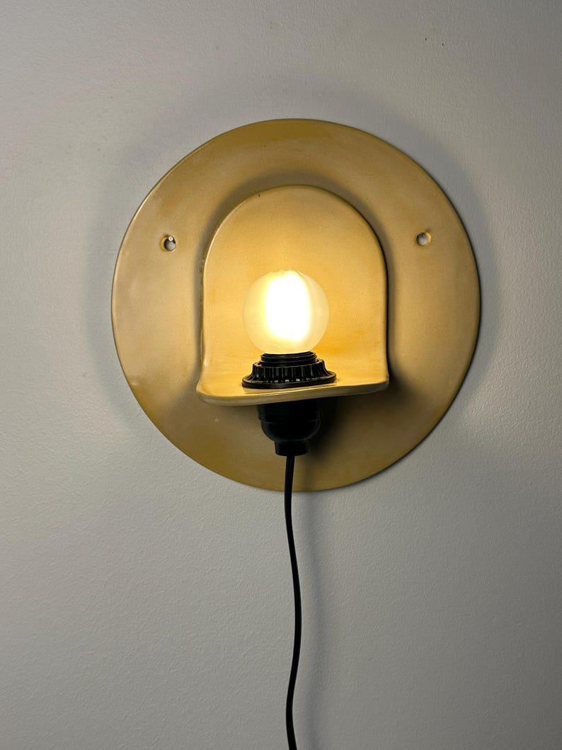 Pu&ograve; includere: Un'applique circolare color giallo senape con una sporgenza centrale ad arco che sostiene una lampadina smerigliata luminosa. L'apparecchio include un portalampada nero e un cavo di alimentazione nero, con due piccoli fori di montaggio.
