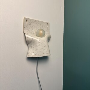Może przedstawiać: Ścienna lampa ceramiczna o białej powierzchni z czarnymi kropkami. Oprawa ma zakrzywiony, pofałdowany kształt z centralnym okrągłym otworem, w kt&oacute;rym znajduje się okrągła, matowa żar&oacute;wka. Poniżej zwisa cienki przew&oacute;d zasilający w jasnym kolorze.
