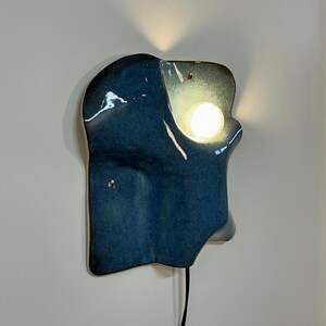 May include: A wall-mounted ceramic sconce with a glossy, deep blue glaze and an irregular, organic shape. The fixture features a circular cutout that emits a warm, glowing light from an integrated bulb, with a black power cord extending downward.