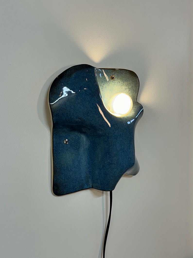 May include: A wall-mounted ceramic sconce with a glossy, deep blue glaze and an irregular, organic shape. The fixture features a circular cutout that emits a warm, glowing light from an integrated bulb, with a black power cord extending downward.