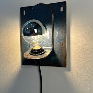 May include: A wall-mounted lamp featuring a rectangular blue metal backplate with a curved shelf. A black socket holds a clear light bulb with a reflective silver-dipped top half. A black power cord extends downward from the base of the light fixture.