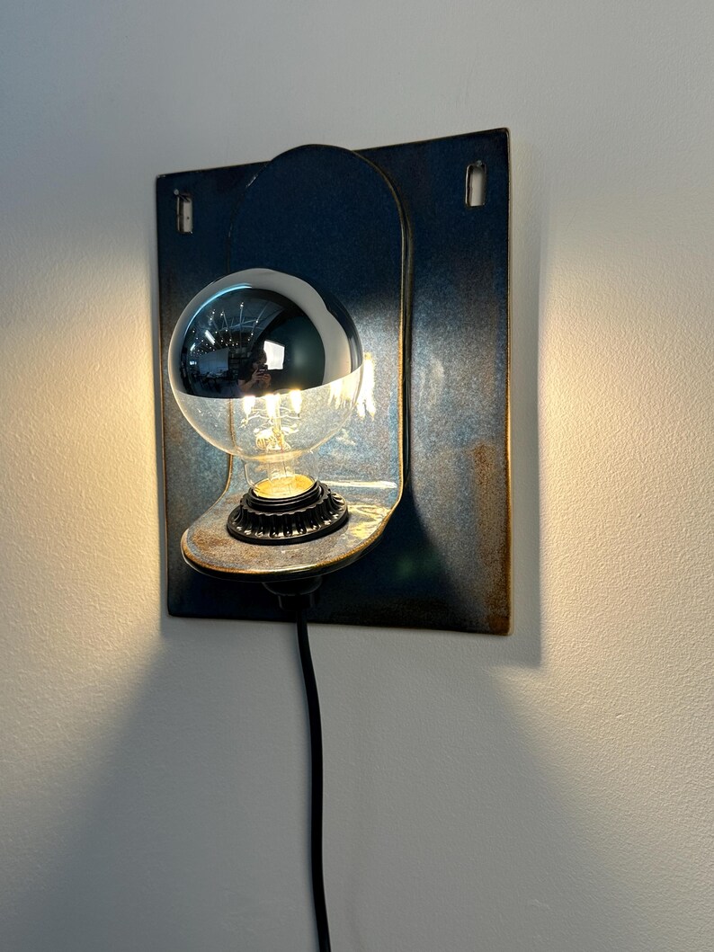 May include: A wall-mounted lamp featuring a rectangular blue metal backplate with a curved shelf. A black socket holds a clear light bulb with a reflective silver-dipped top half. A black power cord extends downward from the base of the light fixture.