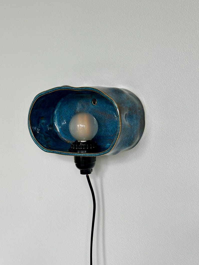 May include: A blue ceramic wall sconce with a bulb. The sconce has a unique, organic shape with a dark blue interior and a lighter blue exterior. A black cord extends from the base of the light fixture.