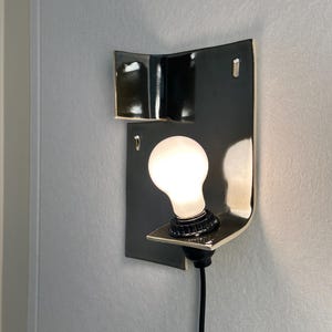 Pode incluir: Arandela de parede moderna com estrutura em metal preto e design curvo. Apresenta uma l&acirc;mpada branca e um cabo preto. A arandela est&aacute; montada numa parede cinza claro. O design &eacute; minimalista e contempor&acirc;neo.
