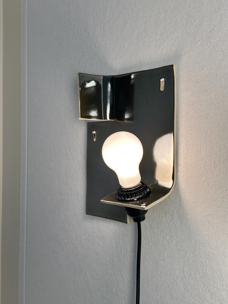 Pode incluir: Arandela de parede moderna com estrutura em metal preto e design curvo. Apresenta uma l&acirc;mpada branca e um cabo preto. A arandela est&aacute; montada numa parede cinza claro. O design &eacute; minimalista e contempor&acirc;neo.