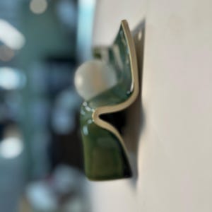 May include: A wall-mounted light fixture featuring a curved, glossy dark green ceramic base with a smooth, off-white finish on the edges. A single, spherical white glass globe bulb is positioned within the wave-like structure of the ceramic form.