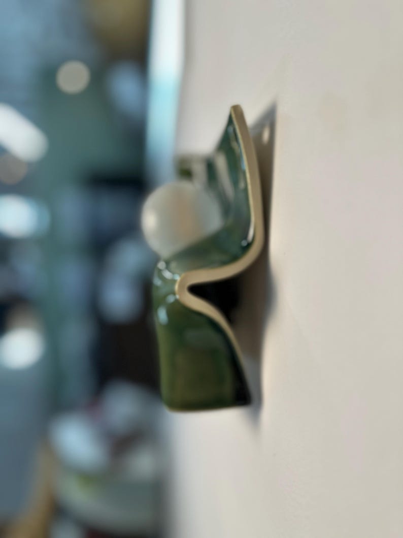 May include: A wall-mounted light fixture featuring a curved, glossy dark green ceramic base with a smooth, off-white finish on the edges. A single, spherical white glass globe bulb is positioned within the wave-like structure of the ceramic form.