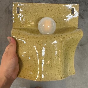 May include: A wall-mounted ceramic light fixture with a glossy, speckled mustard yellow glaze. The piece features a curved, wave-like form with two rectangular mounting holes at the top and a single, central white spherical light bulb.